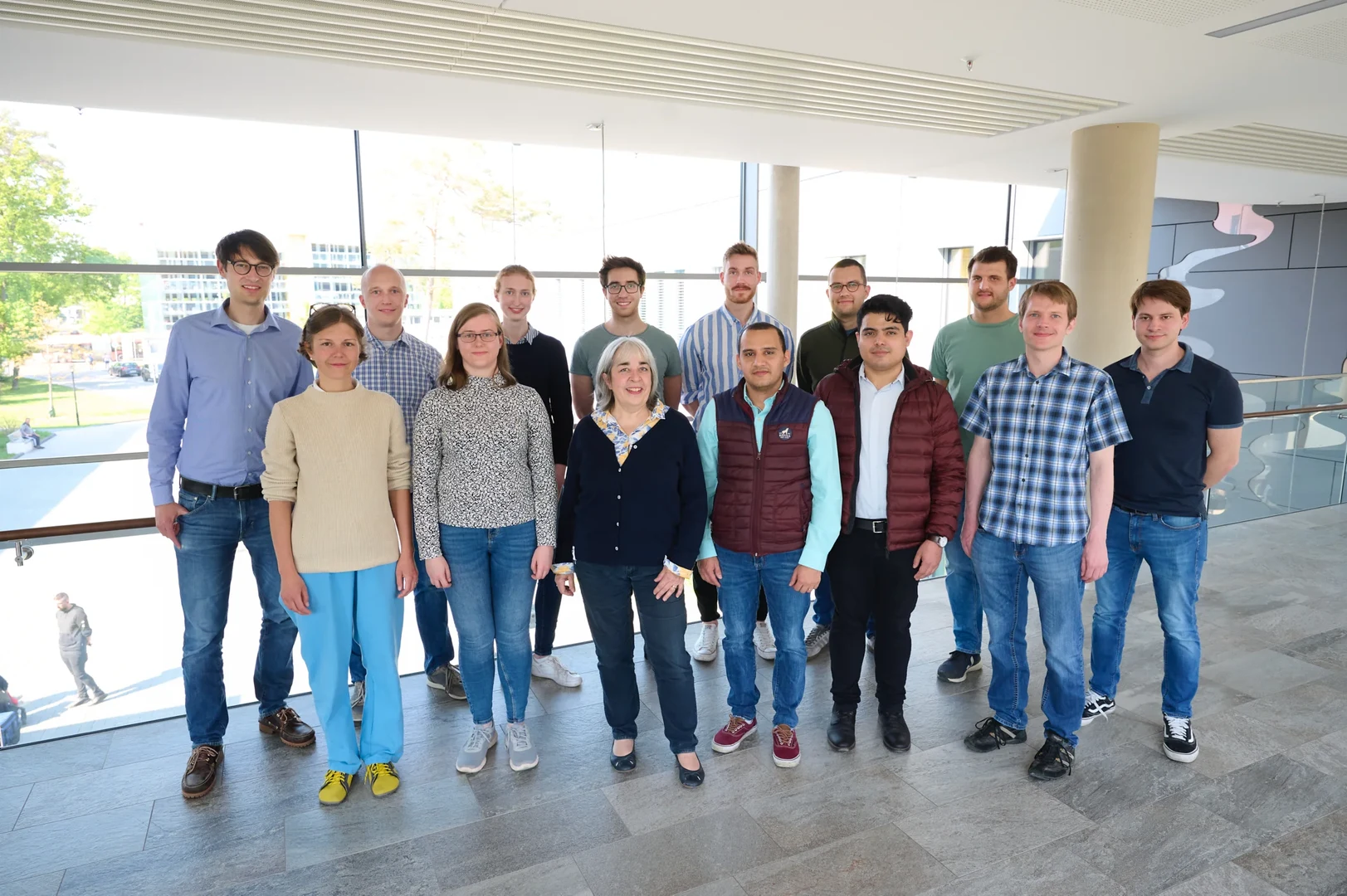 Eine Wissenschaftlerin und ein Wissenschaftler arbeiten hinter einer Glasfassade und mischen Chemikalien mit Großgeräten.